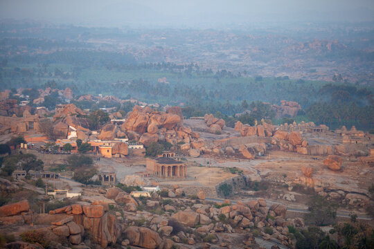 An expansive aerial view of the ancient city of Hampi, India, showcasing a unique landscape dotted with massive boulders, historical ruins, and traditional dwellings amidst lush greenery under a soft, - Powered by Adobe