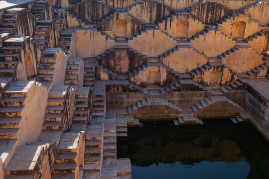 The mesmerizing geometric patterns of a historic Indian stepwell, with countless ancient stairs descending to a dark pool of water, revealing an engineering marvel and a profound sense of history and - Powered by Adobe