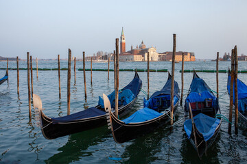 Iconic Venetian gondolas moored in the tranquil lagoon with the stunning San Giorgio Maggiore Basilica and its bell tower visible in the background under clear skies, embodying timeless romance and Eu