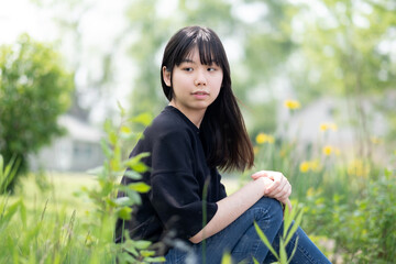 Young asian teenage girl sitting on the ground in a grassy area, surrounded by green plants and wildflowers, wearing a black top and jeans.