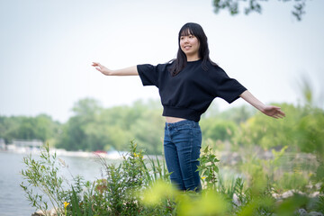 Young asian teenage girl standing on the ground in a grassy area, surrounded by green plants and wildflowers, wearing a black top and jeans.