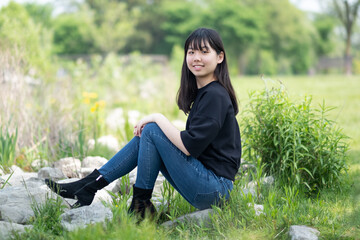 Young asian teenage girl sitting on the ground in a grassy area, surrounded by green plants and wildflowers, wearing a black top and jeans.