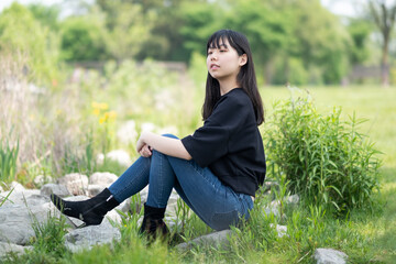 Young asian teenage girl sitting on the ground in a grassy area, surrounded by green plants and wildflowers, wearing a black top and jeans.