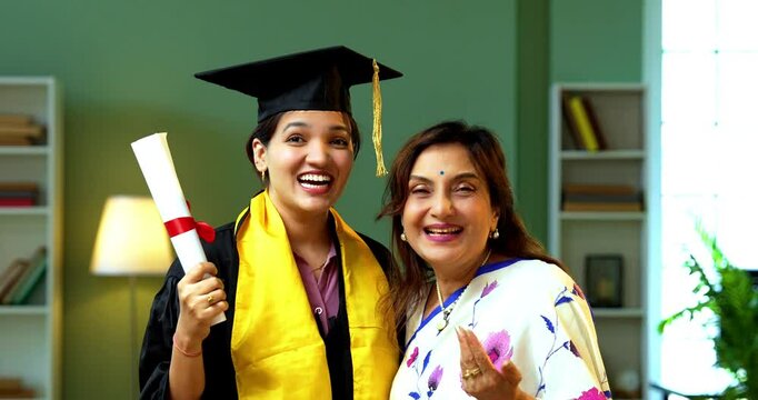 Indian Happy female graduation student with her mother, Indian asian young girl proudly showing graduation degree with gown, hat to senior mom at home while both celebrating success hugging with joy