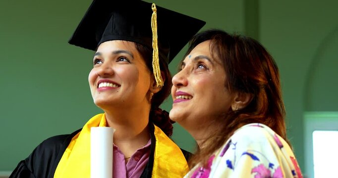 Indian Happy female graduation student with her mother, Indian asian young girl proudly showing graduation degree with gown, hat to senior mom at home while both celebrating success hugging with joy