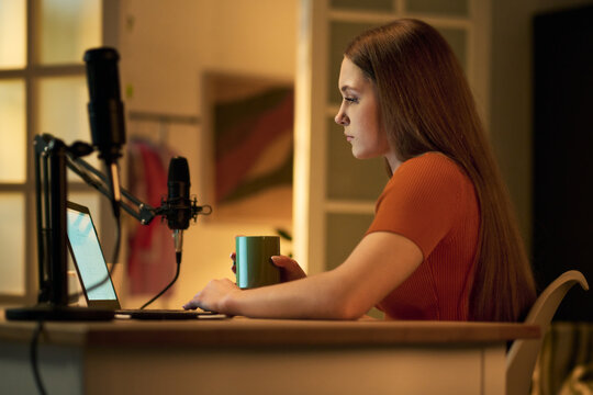 Caucasian teenage girl sitting at desk recording podcast using professional microphone and laptop, holding mug in hand, focused on screen, modern home interior visible in background
