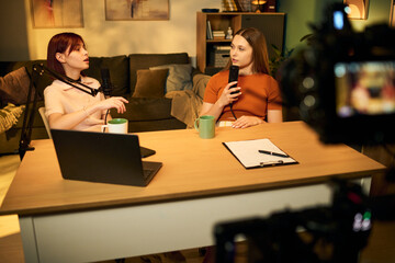 Two Caucasian teenage girls recording podcast in studio, sitting at table with microphones, laptop and clipboard, engaging in conversation while being filmed by camera
