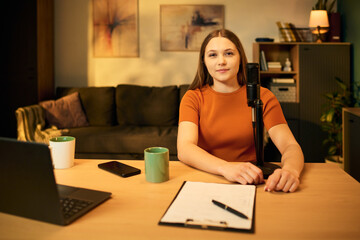 Portrait of Caucasian teenage girl sitting at desk recording podcast using professional microphone...
