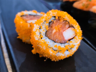Sushi rolls topped with vibrant orange tobiko, filled with salmon and crab stick, served on a black ceramic plate in Japanese restaurant setting