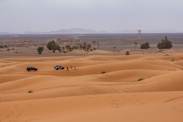 Expansive golden sand dunes in the foreground give way to a distant oasis village with palm trees and off-road vehicles, showcasing desert exploration
