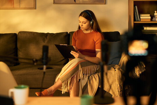 Caucasian teenage girl sitting on sofa wearing headphones preparing podcast notes on clipboard with professional microphone and camera equipment visible in foreground