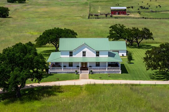 Aerial View of a White Farmhouse with Green Metal Roof Surrounded by Lush Green Fields and Mature Trees a Red Barn and Cattle are Visible in the Background