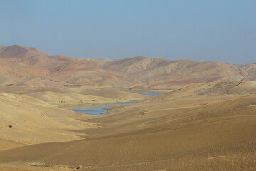 An expansive, arid mountain landscape features rolling hills with a large, tranquil blue lake nestled in a valley, under a clear blue sky, depicting a serene and vast natural environment.