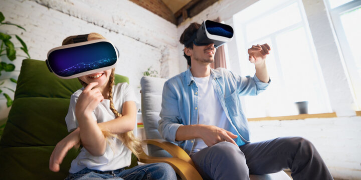 A father and his daughter, both Caucasian, enjoy a virtual reality headset experience at home. Concept of family bonding through technology and immersive experiences.