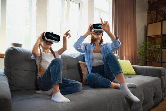 A Caucasian mother and daughter are enjoying a virtual reality experience together on a cozy living room sofa. Concept of family bonding through technology.