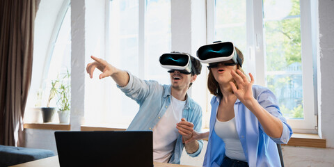 A Caucasian man and woman are engaged in a virtual reality adventure, wearing headsets as they...