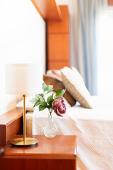 Detail of a master bedroom nightstand. Lamp with a round, gold base, round lampshade, white lampshade, glass vase, white plaid bedspread, wooden furniture, blue curtain, bed