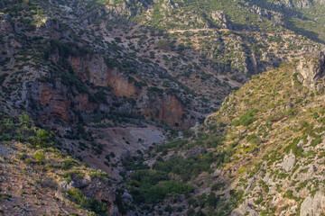 A dramatic, rocky gorge with steep, arid slopes and sparse vegetation descends into a valley, revealing a rugged and challenging natural landscape.