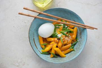 Korean tteokbokki served in curry sauce with boiled egg in a turquoise bowl, horizontal shot on a light-beige stone background, elevated view
