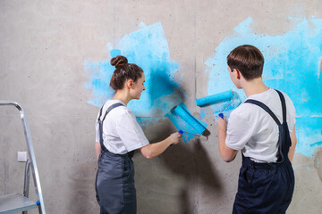 House decoration DIY renovation concept. Happy couple in empty room of new home painting wall together. Husband and wife holding paint roller painting wall with blue color paint in new house