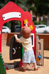 A toddler wearing a sun hat and summer clothes is engaged at an outdoor playground. The child is interacting with a colourful play structure featuring a red panel. Lifestyle photo, summer time.
