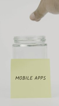 A yellow sticky note with the words mobile apps is attached to a glass jar as a hand inserts a coin. This vertical video reflects saving for digital tools and mobile app expenses