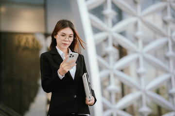 Fototapeta premium Businesswoman in black suit holding laptop and smartphone smiling confidently outdoors in modern urban environment