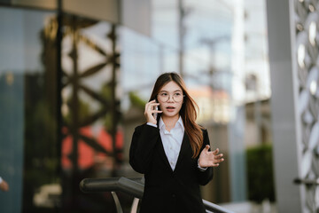 Young businesswoman wearing glasses and a black suit holds coffee and smartphone while smiling confidently outdoors in a modern urban setting.