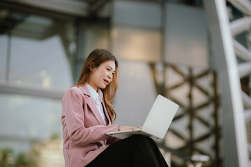 Fototapeta premium Young businesswoman in a pink blazer using a laptop outdoors, standing confidently near a modern office building with decorative glass facade.
