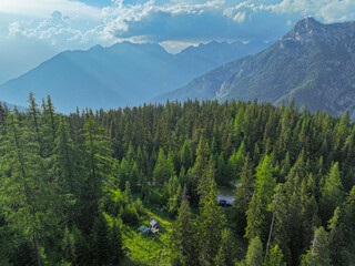 AERIAL: Discreet campsite nestled within a dense evergreen forest, with a car parked nearby and majestic mountains rising under a partially cloudy sky. Outdoor adventure in pristine alpine nature.