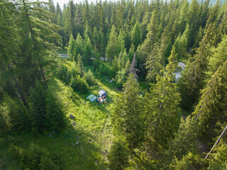 AERIAL: Secluded wild campsite with two tents and camping gear amidst tall green trees. Sunlight...