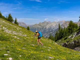 Young woman with a large blue backpack and trekking poles ascends a steep, grassy mountain slope...