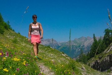 PORTRAIT: Woman with a backpack and sunglasses walks uphill on a sunlit mountain trail. Path is...