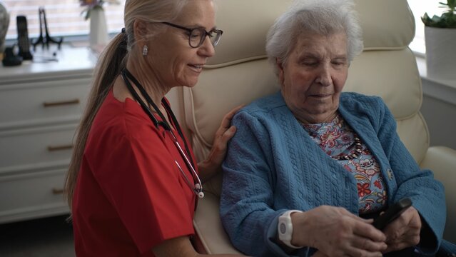 A healthcare worker guides an elderly woman in using her remote at a home setting. The interaction highlights technology support for seniors while sitting comfortably.