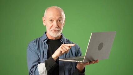 Portrait of senior entrepreneur gesturing while holding a laptop with a green screen backdrop in a studio. He showcases confidence and professionalism in digital work.
