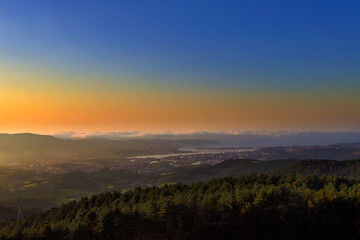 Obraz premium Colorful sunrise over city and sea of fog covering valley and hills with forest in foreground
