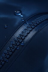 Macro close-up of a waterproof zipper with thick interlocking plastic teeth on dark blue technical fabric. Water droplets are scattered across both the textile and the zipper surface