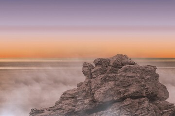Rugged rock formation is emerging from calm sea in flat design, featuring misty waves, gradient sky