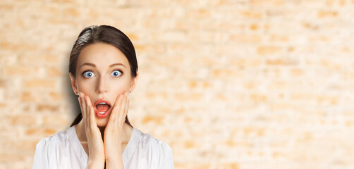Wow! Excited surprise, expressive happy black hair woman in white shirt. Funny girl with wide open eyes, mouth, hands on cheeks. Shop sales, discounts - ad concept banner image. Bricks wall background