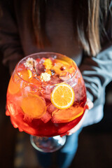 Close-up of a person holding a large glass bowl filled with red summer punch, ice cubes, orange...