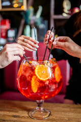 Four people share a large vibrant cocktail with straws in a cozy bar. Garnished with orange slices and flowers, the drink evokes a festive and social atmosphere.
