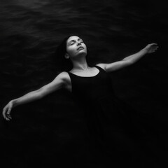 Serene woman in a black swimsuit floats on her back in dark water, eyes closed. Peaceful, minimalist black and white image evoking tranquility.