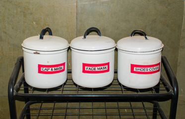 iron rack placed outside an ICU in a hospital, containing three white tin boxes labeled Cap and Mask, Face Mask, and Shoe Cover. reflects hospital hygiene protocols and infection control measures.