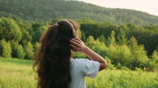 Cheerful teenage girl listening to music on headphones and enjoying an incredible warm sunset on background amazing mountains. Childhood and summer weekend concept