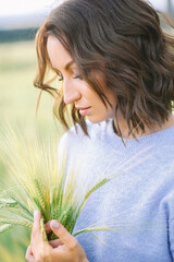 A young woman with brown hair holds a wheat ear in her hands, looking at it thoughtfully. The...