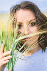 A young woman with brown hair, in a blue sweater, holds stalks of wheat. The look is expressive,...