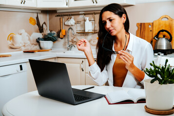 Woman studying online at home kitchen, taking an online course, holding glasses thoughtfully, with...
