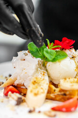 Close-up of a chef's gloved hand sprinkling seasoning over a gourmet dessert with banana slices,...