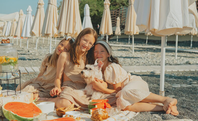 Three happy girls hugging a small white dog at a beach picnic with watermelon and lemonade on a...