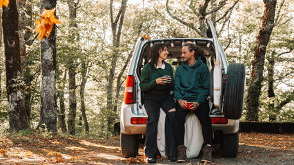 Obraz premium Young couple sitting in trunk, drinking hot beverages and enjoying peaceful autumn forest together